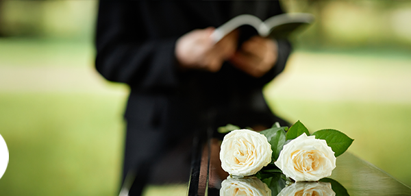 Close up on white roses on a casket outdoors with person dressed in black holding a back in the blurred background