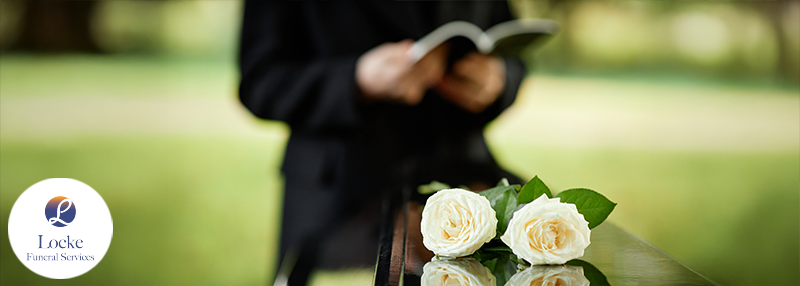 Close up on white roses on a casket outdoors with person dressed in black holding a back in the blurred background