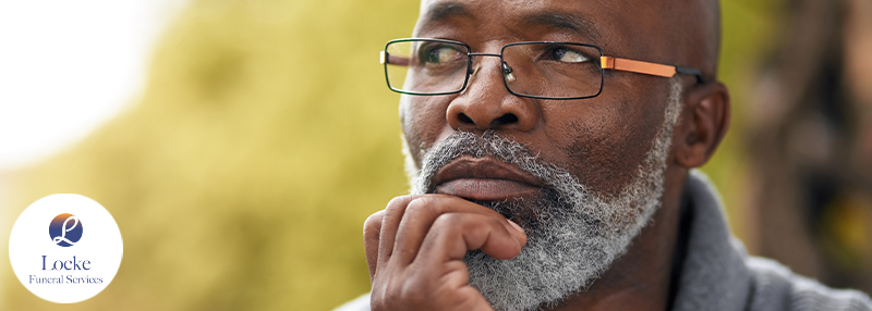 Image of man with glasses on sitting outside and holding his hand to his beard thinking