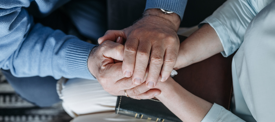 Person in green sweater holding the hands of another person in blue sweater with hands over a Bible